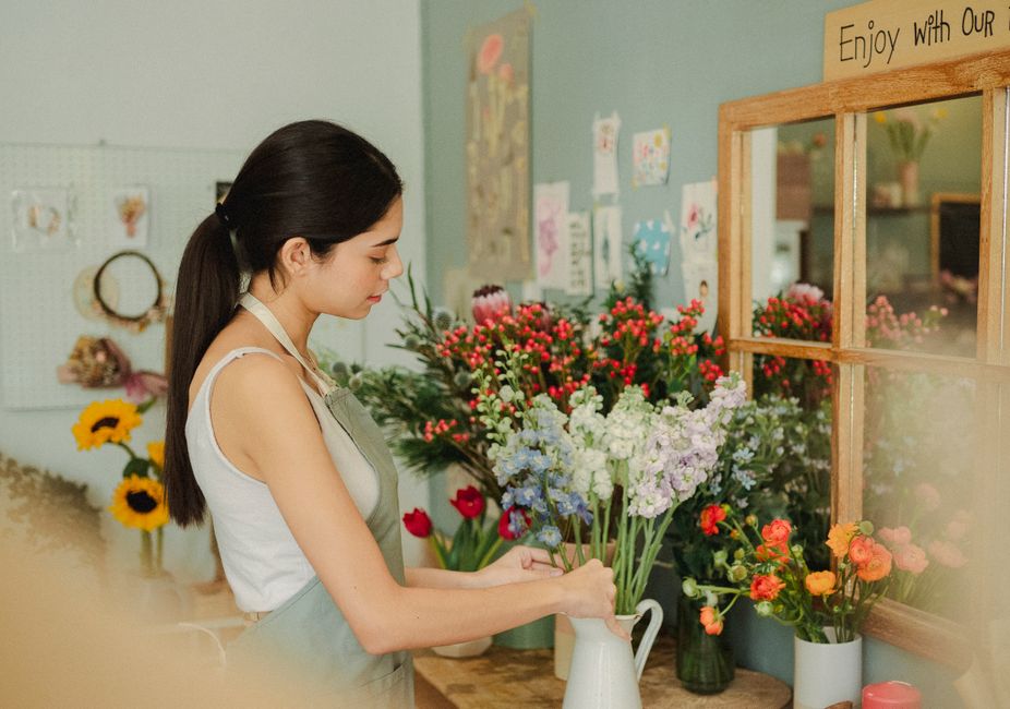 A young woman with dark hair tied back, wearing a white sleeveless top and a grey apron, arranging a bouquet of mixed fresh flowers inside a florist shop. The bouquet includes light blue, white, and p