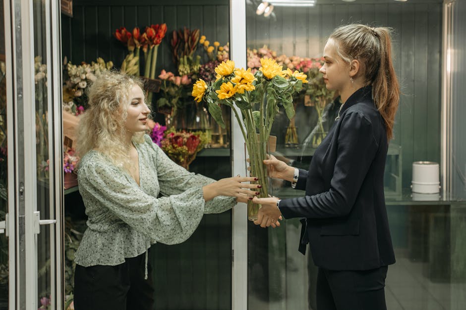 A florist with curly blonde hair, wearing a light gray floral blouse, hands over a bouquet of bright yellow sunflowers with green stems and leaves to a customer inside a flower shop. The customer has 