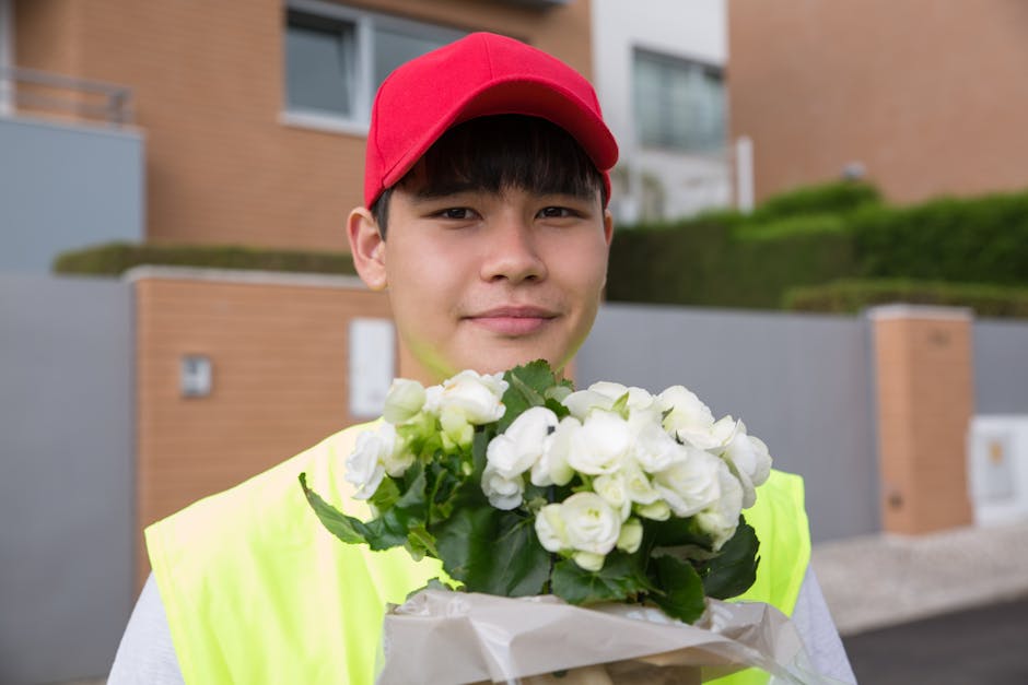 A young male flower delivery courier wearing a red cap and a high-visibility yellow vest is holding a bouquet of fresh white roses with green foliage wrapped in white paper. He is standing outdoors in